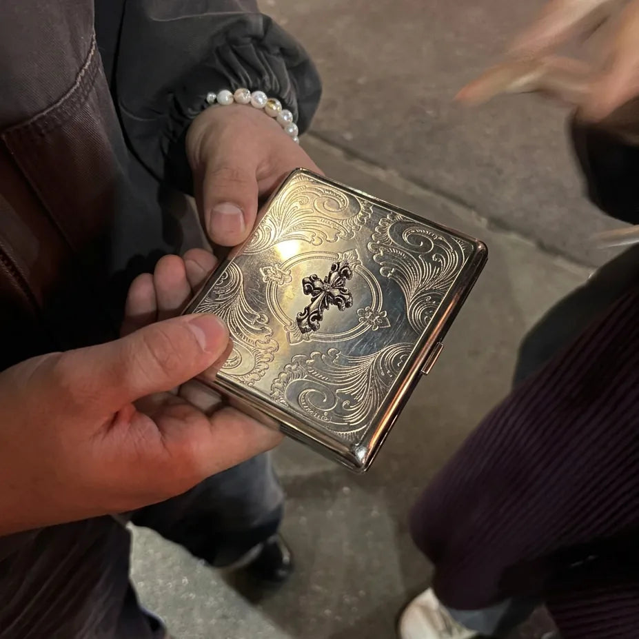 Hands holding an engraved silver tin card case with a gothic cross, on a city sidewalk